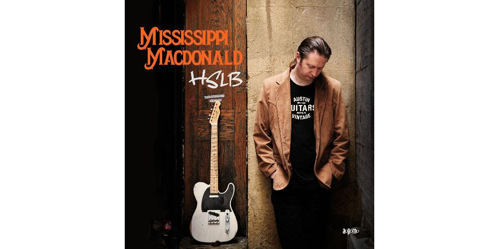 A man standing next to a guitar, holding the Mississippi MacDonald CD.