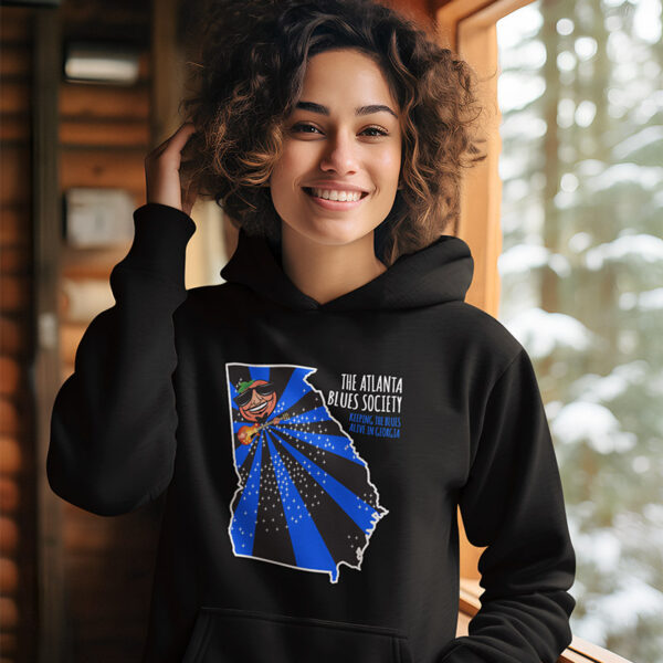 A smiling woman wearing a hoodie with "the atlanta blues society" graphic, standing indoors with a view of trees through the window.