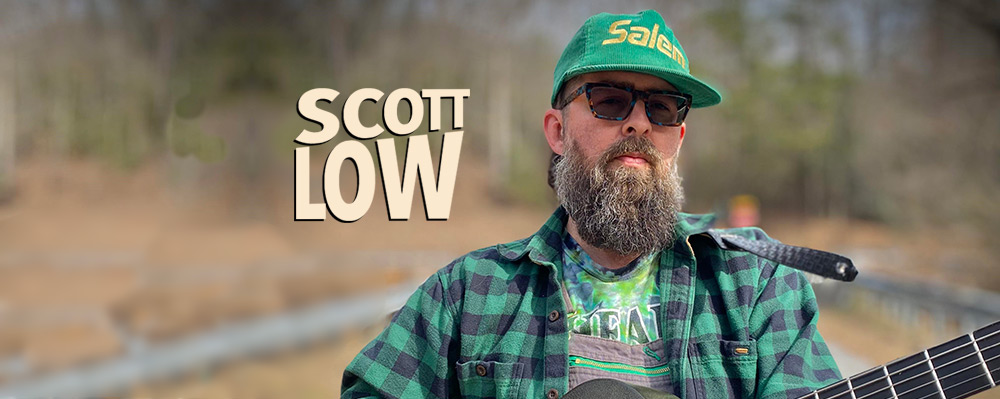 A bearded man wearing a green cap and a flannel shirt holding a guitar with the name "scott low" written above him.