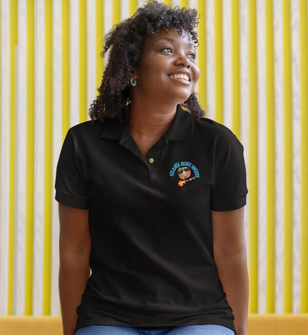 A smiling woman wearing a black polo shirt with a logo on the chest, sitting against a yellow striped background.
