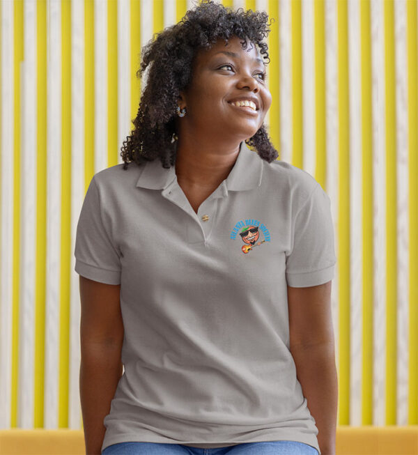 Woman smiling in a grey polo shirt with an embroidered logo, seated against a yellow striped background.